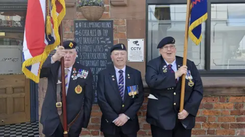 Mitch Cottle Three veterans in dark coloured suits with a number of medals and wearing berets with military pin badges stand outside the pub. Two of them hold military flags.