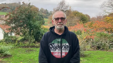 Ian Edmonds wearing a dark hoodie. He has glasses on and a short beard. He is standing on the edge of a small clearing, and you can see Dunster Castle in the background, with a tower just poking over some trees.
