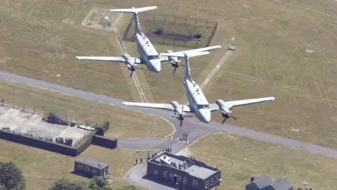 Two Shadow F1 planes fly over buildings, tarmac and green grass at RAF Waddington in Lincolnshire