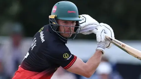Ben Cox, wearing Leicestershire Foxes' black and red shirt and green helmet, plays a shot during the One-Day Cup game against Gloucestershire