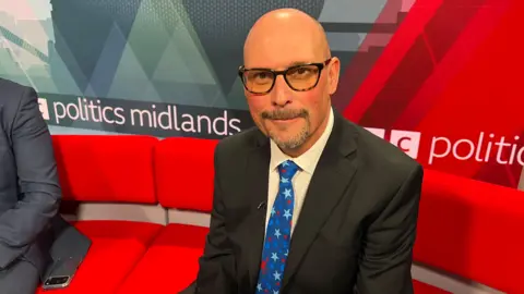 Adam Kent is bald and wears black glasses with yellow tinted lenses. He is wearing a black suit jacket, white shirt and blue tie with light blue and red stars on it. He is sitting on a red sofa with a backdrop behind him that reads "politics midlands"