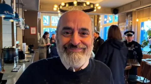 BBC/Chris McHugh Steve Hudson smiles for the camera. He has a bald head and a bushy white beard, wearing a blue jumper over a pink shirt. He is in a pub leaning against a bar with people seen behind him