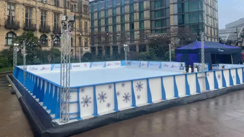 A large ice rink with blue and white barriers sits below tall city centre buildings