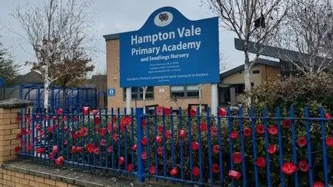 Blue and white-coloured sign saying "Hampton Vale Primary Academy" with a large brick-building in the background with a blue metal fence and red poppies in the foreground.