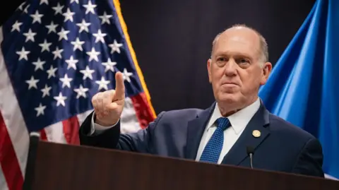 Tom Homan, White House border czar, stands in front of an American flag wearing a blue suit, white shirt and blue tie