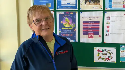 Fran Tinkler, a woman with short blonde hair with a fringe and glasses. She wears a navy coat that reads Manx Mencap and stands in front of a notice board with colourful posters of activities that are taking place.