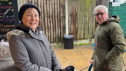 Shaun Whitmore/BBC A man and a woman standing outside on brown stone slabs, with the woman to the the left of the image leaning with her elbow on the metal counter of a food and drink stall. She is wearing a grey coat, black gloves and a black hat and is smiling at the camera. The man to her right is wearing a dark green coat, his left hand in the pocket and his other hand holding a black and white dog on a lead. He has grey hair and tinted glasses.