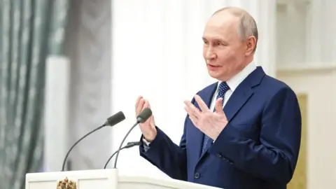 President Putin wearing a white shirt, dotted blue tie and blue suit, gestures with his hands as he speaks at a white podium with two microphones. A light green patterned curtain stands to the left of the picture.