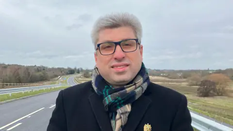 A man with a neutral expression standing in the middle of a single carriageway. He has white hair, black spectacles and is wearing a long black coat with a green and chequered scarf. 