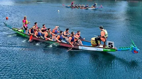 Ten people and two people at each end of the boat are in the water alongside large office buildings in Salford Quay. Three other boats can be seen in the background.