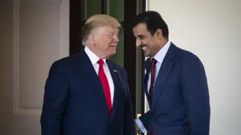 Donald Trump speaks to Tamim bin Hamad Al Thani, who smiles in response. Both men are wearing blue suits and standing outside the White House. 