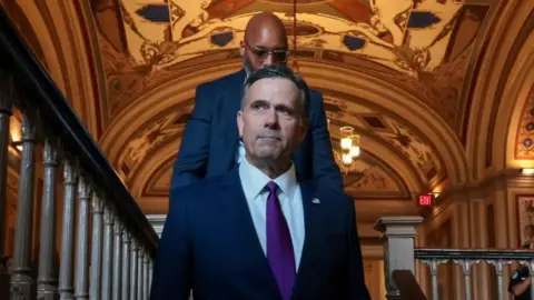 CIA director John Ratcliffe walks down a flight of stairs. A man with a bald head and glasses walks behind him. 