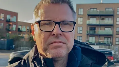 Stephen Huntley/BBC Andy Long wears black-rimmed glasses and is wearing a black coat. He has a sombre expression. He is standing in a car park.