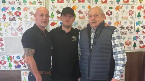 Family handout Three men smiling at the camera. They are inside and behind them is a wall covered in children's drawings. 