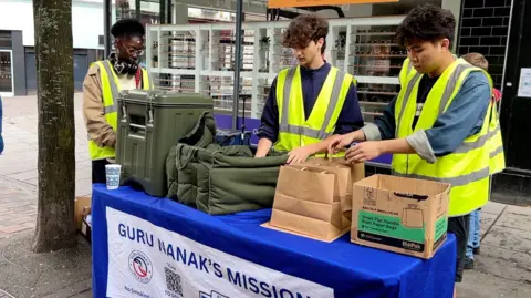 Three volunteers in high vis jackets setting up a food distribution table. Table has large green urn and bags and boxes