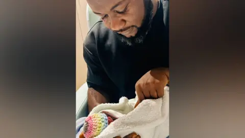 Daniel Udoeyo Daniel is wearing a black t-shirt holding baby Elkan who is wearing a rainbow hat and wrapped in a white blanket.
