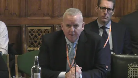 UK Parliament Alistair Macrow wearing a suit and orange lanyard, sitting down speaking to MPs