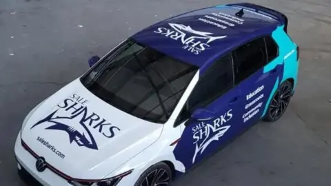 A blue and white Volkswagen Golf car, wrapped in colours and badge of Sale Sharks rugby club