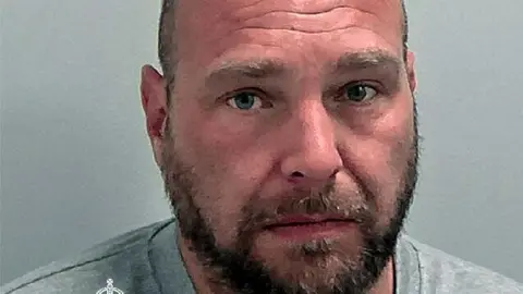 A police photo of a man looking at the camera. He has a brown beard and very short hair and is wearing a grey T-shirt. 