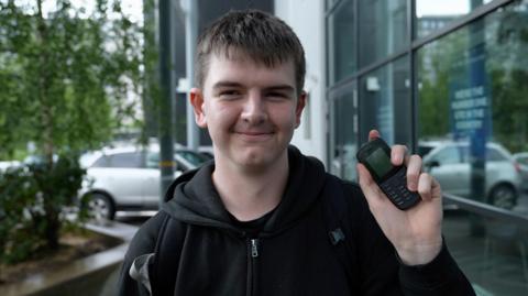 Will, 15, wears a black hoodie and smiles at the camera as he holds up his basic Nokia phone