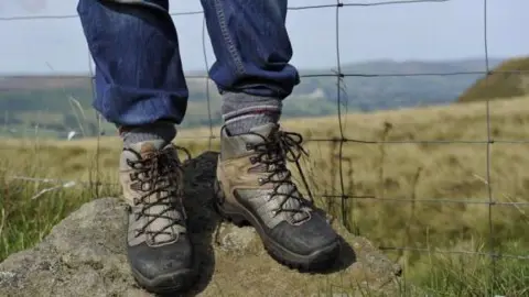 A man wearing walking boots stands on a rock with a wire fence behind him in open countryside.