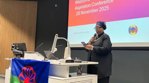 Jon Wright/BBC Pat Bruce-Browne stands in front of a large screen in a lecture theatre. There is a Romani Flag on the front of the desk, a red wheel against a green and blue background. 