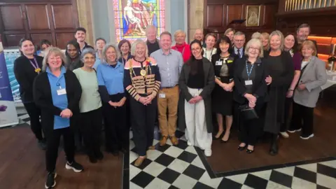 A group of adults, volunteers and clinical staff stand together in a large room posing for the camera