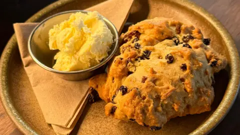 Nettle cafe A crumbly baked good with dried fruit on a plate next to small bowl of clotted cream.