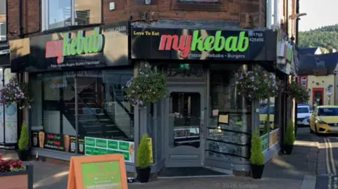 Google A kebab shop is pictured. It is on the corner of two streets and has black branding with red and green writing. The shop is called MyKebab.
