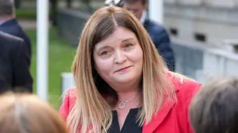 A woman looking at the camera wearing a red top and dark blouse.