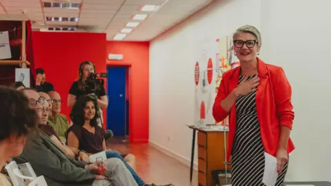 People are listening to a talk by Heather Thomas, who is standing and in the middle of speech, holding a piece of paper. Somebody is filming the talk in the background.