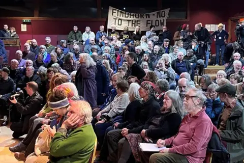 People are sat on seats inside an auditorium-like space. There is a banner at the back of the room with the slogan: "Findhorn - No to the flow".
