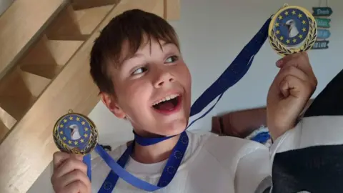 Family photo Cooper, who has short dark blond hair and wears a white T-shirt and part of a gull costume, smiles as he looks at one of two identical medals. The medals have a picture of a seagull on them, surrounded by gold stars on a blue background, and blue ribbons with the EU logo on them.