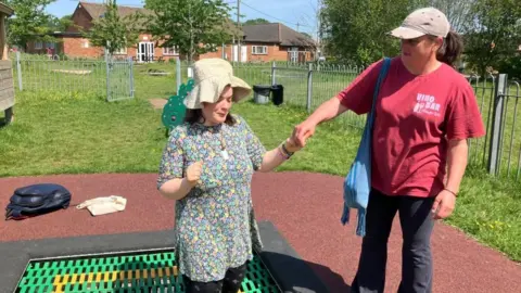 Alison Baker A young woman is at a play park. She is wearing a sun hat and looks happy. A woman is standing next to her, holding her hand. It's a sunny day and both women look happy.