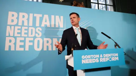 Matt Goodwin stands at a podium which reads "Gorton & Denton needs Reform"