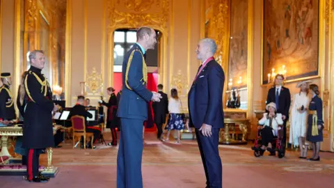 PA Media The Prince of Wales and Graeme Souness are standing facing each other inside Windsor Castle. They are talking and laughing, while several other people mill around in the background. Both men are dressed in suits, and a CBE is hanging around the neck of Souness.