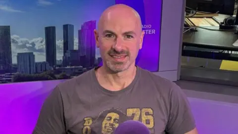 James Goodall, who is wearing a Bob Marley T-shirt, is sitting in the BBC Radio Manchester studio. There is a BBC-branded purple microphone on a desk in front of him. 