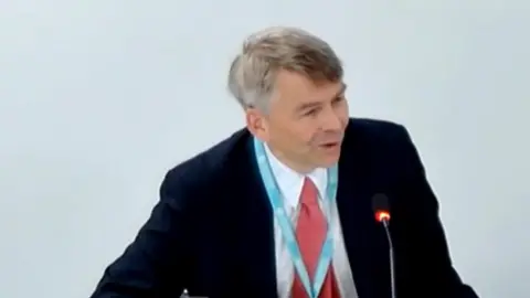 Andrew Gant is sitting at a table at the meeting wearing a dark suit jacket, a pink tie and a white shirt. He is also wearing a blue lanyard around his neck. He has short grey hair and is talking into a microphone.