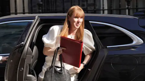 Angela Rayner steps out of a black car wearing a white dress. She carries a red folder and a black bag.