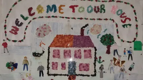 A piece of art which says 'Welcome to Our House' with small figures, a tree and a house below