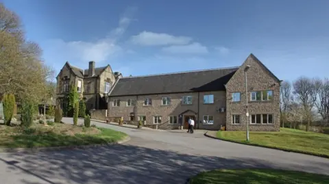 External view of Cygnet Hospital Wyke, with a large driveway, grass verges and trees in the foreground.