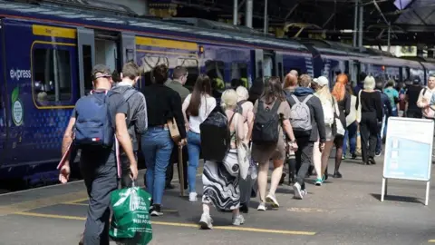 PA Media Passengers queue along a railway platform to board a blue ScotRail train at a busy station, with many carrying backpacks and shopping bags under a covered concourse.