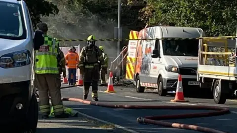 Firefighters and a Cadent gas van can be seen on a street. There are traffic cones on the road.