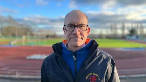 Yeovil Olympiads Athletic Club chairman Geoff Cole stood next to the athetics track in a blue coat, looking directly to camera