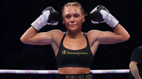 Tiah -Mai Ayton flexing her biceps while looking into the camera. She is standing in the boxing ring wearing a black sports bra, black boxing gloves with her hair plaited into tight cornrows. 