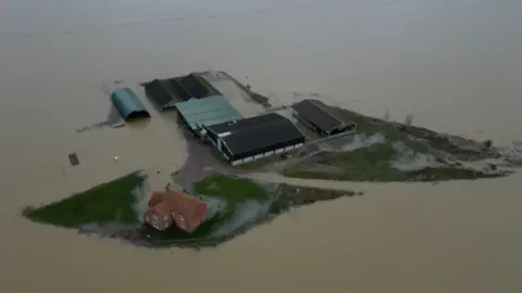 A farmhouse, completely surrounded by water. Some of the farm buildings stand on dry land, but the fields are completely submerged.
