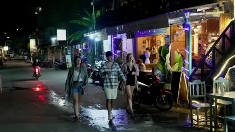 A group of three young people are walking along a street in Vang Vieng with bars visible in the distance