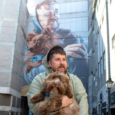 Nuart 2026 A man holding a dog, recreating a large mural on a wall in the background.