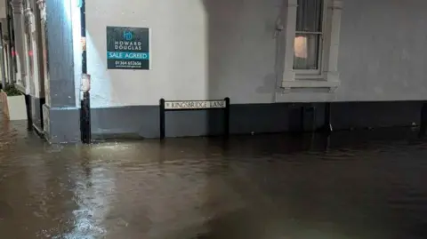 Kate Jones Flood wateron Kingsbridge Lane in Ashburton there is a sign against the building at the centre and a sign on the wall. There is a window on the right.