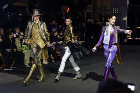 Getty Images 3 models in stride confidently wearing flamboyant outfits along the catwalk